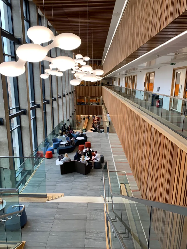 Pohľad zo schodiska na átrium s drevenou fasádou a študentmi pri stoloch na Coventry University