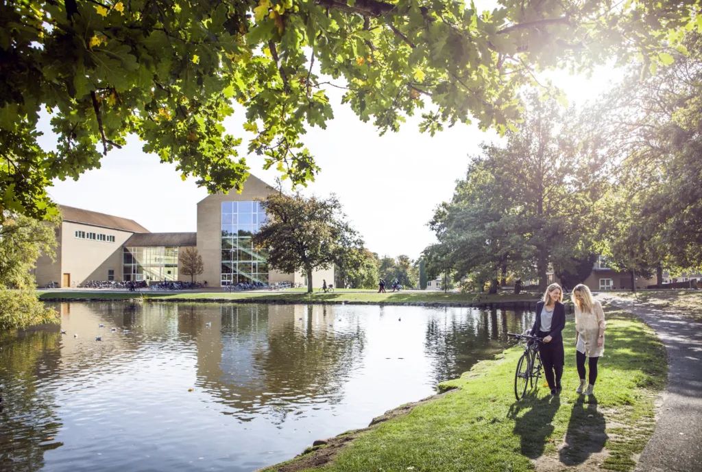 Jazero a budova v univerzitnom parku Aarhus University, dve študentky s bicyklom na brehu